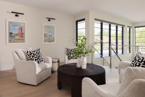 Bright contemporary living room with warm light-wood flooring, off-white walls, three cream upholstered armchairs, and a large round dark coffee table. Black-and-white geometric cushions and framed coastal-style artwork provide contrast. Black-framed floor-to-ceiling windows overlook greenery, while a metal balustrade borders an open stair or landing area.