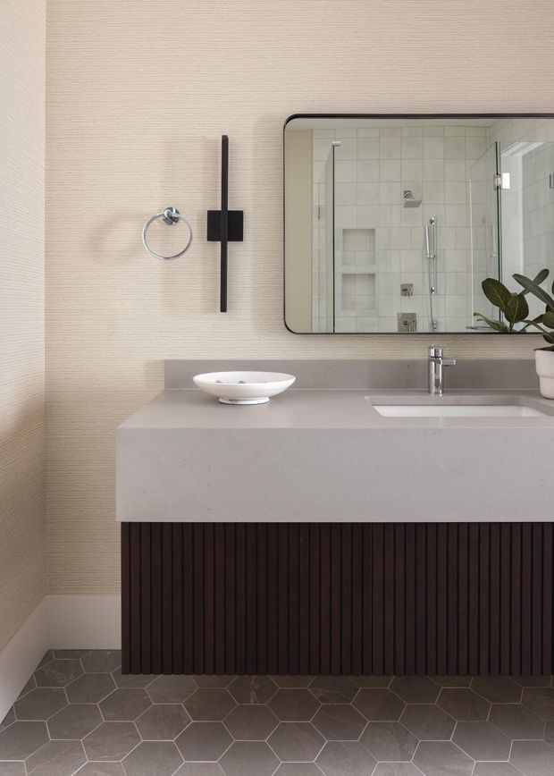 Contemporary bathroom vanity with a floating dark fluted timber-style cabinet, pale solid-surface benchtop, integrated rectangular basin and chrome tapware. A rounded-corner black-framed mirror reflects a tiled shower with recessed niches. The walls have subtle textured beige wallpaper or wall covering, while the floor is finished in dark grey hexagonal tiles. Minimal black wall accessories and a small plant add contrast.