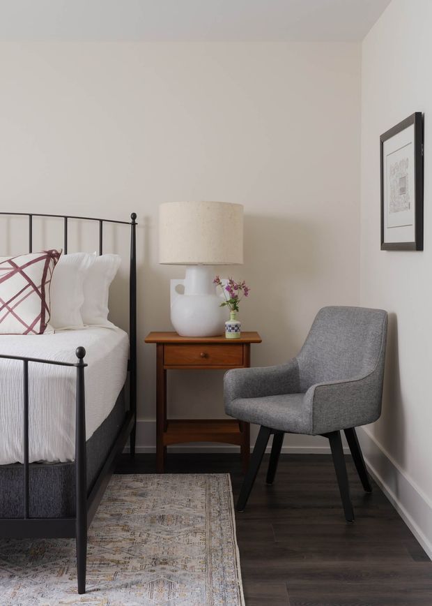 A compact, softly styled bedroom corner featuring a black metal spindle bed with white bedding and a muted patterned accent pillow. Beside it is a small mid-tone timber bedside table with a drawer, topped by a white ceramic lamp with a cylindrical fabric shade and a small vase of flowers. A grey upholstered armchair sits against the pale wall, with framed artwork above it. Dark timber flooring and a neutral patterned rug complete the calm, traditional-contemporary interior.