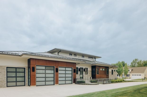 Exterior view of a contemporary low-rise residential property with multiple sectional garage doors, warm wood-toned cladding, pale stone or masonry walls, a shallow metal roof, and a covered entry porch with a ramp and railing. The broad concrete driveway and neighbouring homes are visible under an overcast sky.