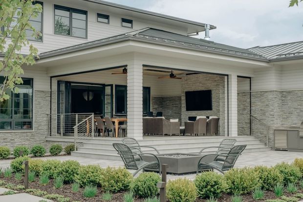Exterior view of a contemporary two-storey home with a covered outdoor entertaining terrace. The terrace includes a dining area, lounge seating, wall-mounted television, outdoor fireplace or feature stone wall, ceiling fans, and a fire table. White horizontal cladding, dark-framed glazing, and a grey metal roof define the facade, with landscaped garden beds in the foreground.