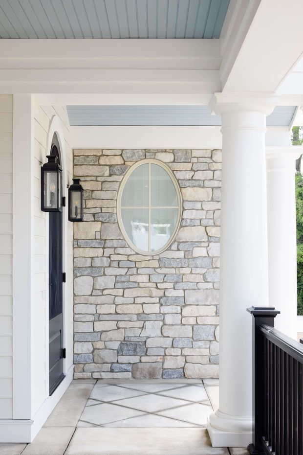 A bright covered residential entry porch featuring white horizontal weatherboard walls, a pale blue beadboard ceiling, substantial white classical columns and beams, dark front door and railings, and a rustic light-grey natural stone accent wall. The stone wall includes a large oval window with pale trim, while the porch floor is laid with large pale stone or concrete slabs in a geometric pattern. Traditional coastal-inspired detailing dominates the facade.