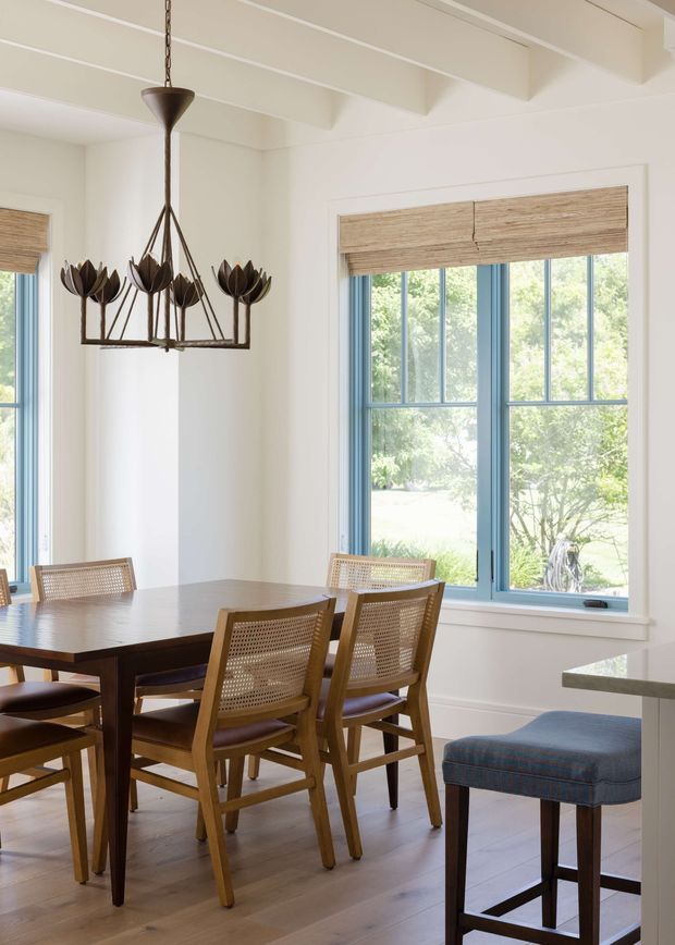Bright dining area with a dark timber dining table and woven cane-back chairs, positioned beside large blue-grey framed windows. The room features white walls, exposed painted ceiling beams, light timber flooring, woven window shades, and a sculptural dark metal floral chandelier. A portion of a kitchen island and upholstered counter stool is visible at right.