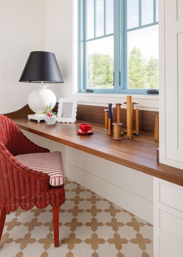 Bright, refined interior corner featuring a built-in timber window seat or desk beneath large pale blue-framed windows. The space includes white cabinetry, a warm dark timber benchtop, patterned geometric floor tiles, a black-and-white table lamp, decorative wooden spindles, and a red woven armchair with striped cushion.
