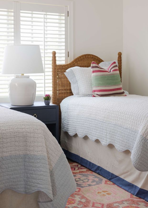 Bright, cozy bedroom featuring two twin beds with woven rattan headboards, white quilted bedding, and a striped multicolour accent pillow. A dark bedside table with a white ceramic lamp sits between the beds. White plantation shutters cover the window, while a patterned pink and blue area rug adds colour beneath the beds.