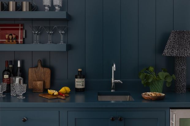 A dark, richly coloured bar or compact kitchen beverage station with deep navy vertical-panel walls, matching open shelves and base cabinetry. The space includes a stainless-steel sink and tap, glassware, wine and spirits bottles, a timber chopping board, citrus on a cutting board, and a small indoor plant. A textured dark lamp sits at the right, adding a moody decorative accent.