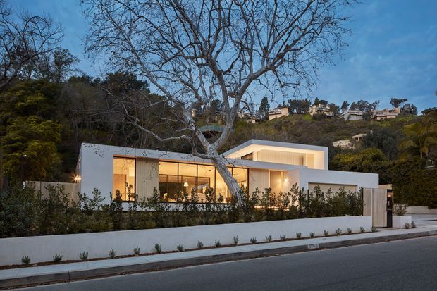 A modern, low-profile white residence viewed from across the street at dusk. The home features crisp geometric volumes, expansive floor-to-ceiling glazing, a projecting upper-level box, and warm interior illumination. It sits behind a low white boundary wall with sparse landscaping, mature trees, and a wooded, gently elevated suburban backdrop.