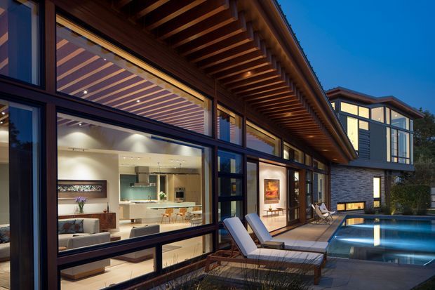 Twilight exterior view of a contemporary residence with extensive floor-to-ceiling glazing, dark framed windows, warm timber soffits and exposed pergola-style roof beams. The open-plan living and kitchen areas are visible through the glazing, while a paved terrace with lounge chairs leads to a rectangular swimming pool. A projecting upper-level wing is visible at the right.