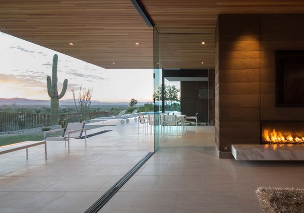 A contemporary indoor-outdoor living area with fully retractable floor-to-ceiling glass walls opening to a pool terrace and expansive desert landscape. The interior features a warm timber-lined ceiling, large-format pale tiled flooring, dark wood wall panelling, and a built-in linear fireplace. A dining area and outdoor seating are visible beyond the glazing.