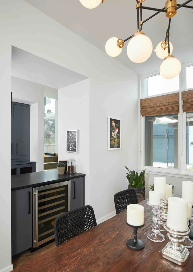Bright dining area with a dark timber dining table, black woven chairs, and a modern multi-globe pendant light. A compact dark built-in drinks cabinet with glass-front wine refrigerator is visible beside the opening to the kitchen. White walls, tall windows with woven timber blinds, indoor plants, and candlelit table styling create a warm, contemporary interior.