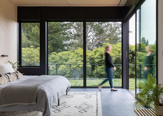 A contemporary bedroom with a low platform bed, textured grey and white bedding, and a patterned area rug. Full-height black-framed glazing opens onto a balcony or terrace overlooking lush greenery, bringing abundant natural light into the room. A large mirror or reflective glass panel on the right repeats the view and the walking figure. The interior has a restrained monochromatic palette with indoor palms and natural outlook.