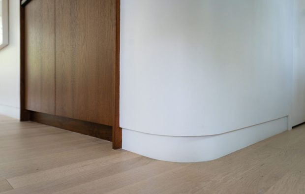 Close-up interior view of a minimalist curved white wall or built-in partition meeting pale timber flooring, with a tall dark timber-clad joinery or doorway element at left. The scene is uncluttered and focused on clean architectural detailing and material transitions.
