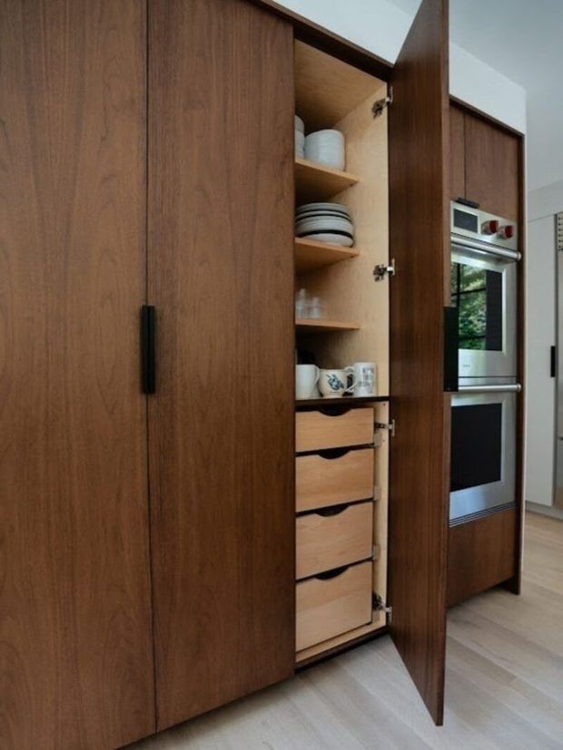 Close-up view of a tall, custom timber kitchen storage wall with an open pantry cabinet. The pantry contains adjustable shelves displaying crockery and glassware, along with several deep drawers below. Adjacent integrated stainless-steel wall ovens are visible within matching dark timber cabinetry. The space has pale timber flooring and a clean, understated contemporary fit-out.