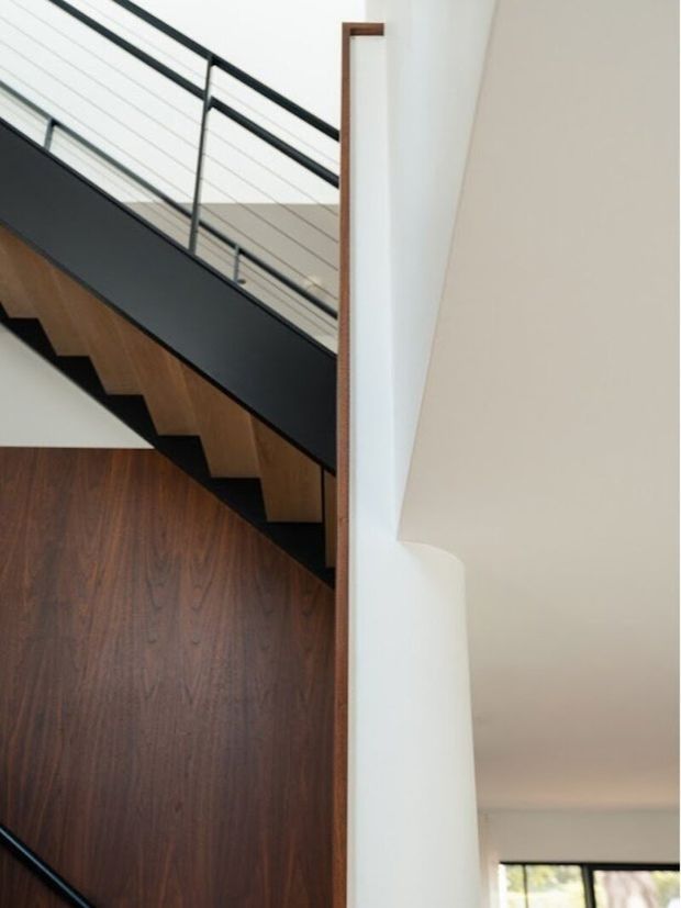 Interior architectural detail showing a modern stairway with dark timber treads and richly toned timber wall or paneling, a black metal stringer, and slim horizontal cable railing at an upper landing. White plaster walls and a tall vertical timber trim create a crisp, high-contrast composition. A small portion of glazing and the adjacent room is visible in the lower right.