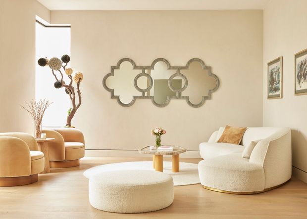 A warm, refined living room with a cream boucle sofa and matching rounded ottoman, two curved lounge chairs by a bright window, a pale timber floor, and a sculptural mirrored wall feature. Neutral beige walls, framed artwork, soft furnishings, and restrained decor create a softly luxurious contemporary interior.