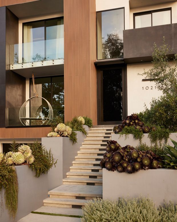 Contemporary multi-storey residential facade viewed from the landscaped front entry. The home features a warm vertical ribbed timber-clad section, smooth light render, dark-framed glazing, a projecting dark balcony, and a sculptural stairway leading to a recessed black front door. Layered raised planters contain drought-tolerant succulents and ornamental planting, while a hanging circular chair is visible behind the lower-level glazing.