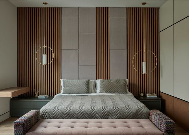 A sophisticated bedroom with a centred upholstered bed, grey textured bedding, and a large tufted bench at the foot. The feature wall combines vertical dark timber battens with pale padded or panelled sections, complemented by dark built-in bedside cabinetry and warm wood wall panelling. Two sculptural pendant lights hang symmetrically over the bedside areas. The room has a refined, moody, contemporary feel with strong geometric composition.