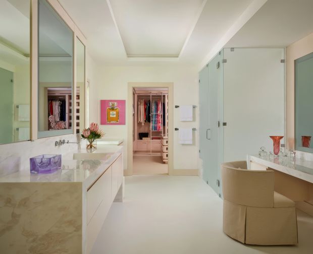 A bright, luxurious dressing room and vanity area with extensive built-in cabinetry, full-height mirrored and frosted-glass wardrobe doors, a pale stone vanity, upholstered chair, and a recessed ceiling detail. An open walk-in wardrobe is visible at the rear, with a clean cream-and-white palette and polished contemporary detailing.