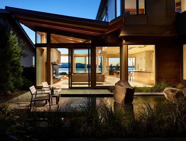 Twilight exterior view of a contemporary multi-level timber-clad house opening onto a landscaped courtyard with a shallow reflecting pool and sculptural stone feature. Extensive floor-to-ceiling glazing reveals a warm living area, dining space and kitchen inside, with views toward a body of water and distant hills.