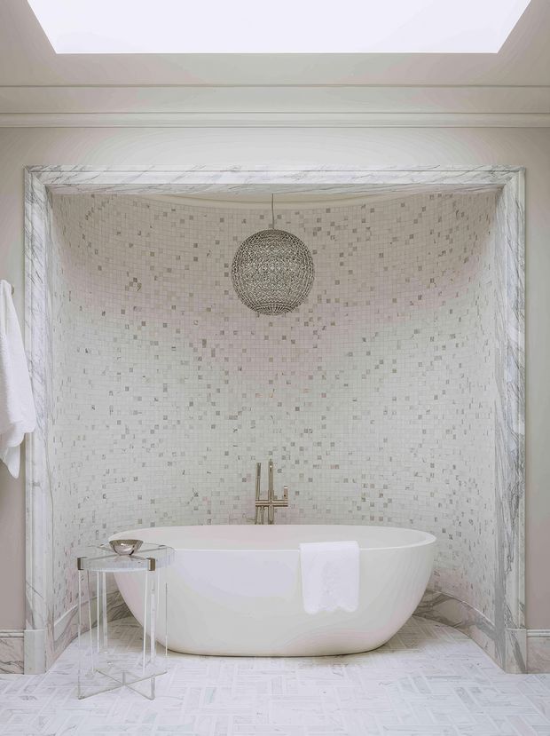 Bright, luxurious bathroom featuring a freestanding white bathtub set within a curved mosaic-tiled alcove. The walls and floor use pale marble-like mosaic finishes, with a brass-toned floor-mounted bath filler, a spherical pendant light, a skylight, and white architectural trim. A small metallic side table sits beside the tub.