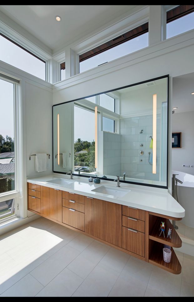 Bright, spacious bathroom with a long double vanity beneath a large black-framed mirror. The vanity has warm timber cabinetry, a white engineered-looking benchtop, integrated rectangular basins, and open curved shelving at one end. White tiled walls and light grey floor tiles create a clean contemporary setting. Large windows and high-level glazing bring in abundant natural light and overlook greenery; a glass-enclosed shower and part of a freestanding bath are visible in the background.