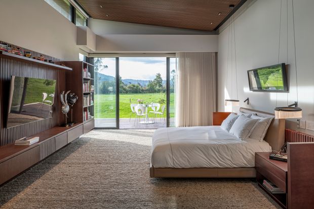 A spacious, refined bedroom with a large bed facing a built-in timber media and shelving wall. Floor-to-ceiling sliding glass doors open to a green rural landscape and outdoor seating. The room features a warm slatted timber ceiling, pale walls, integrated cabinetry, soft neutral bedding, and a textured carpet, with a contemporary built-in desk or bedside unit.