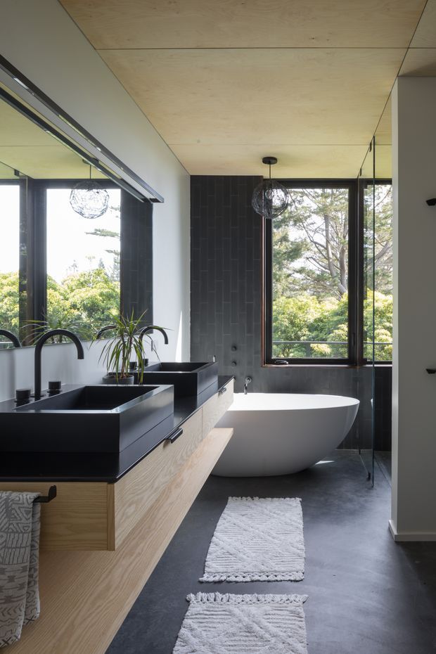 Contemporary bathroom with a freestanding white bathtub positioned beside large black-framed windows, a double dark vanity with sculptural black faucets, and light timber cabinetry. The room features dark tiled walls, a smooth dark floor, a pale timber-lined ceiling, and abundant natural light with leafy views outside.