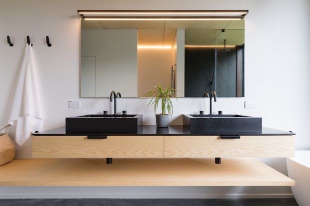 Contemporary double vanity bathroom with a wall-mounted light timber vanity, black stone-look benchtop, two black vessel basins and matching black tapware. A large mirrored cabinet spans the vanity, with warm integrated lighting, white walls, black wall hooks and a potted plant. The room has a clean, refined, high-contrast palette with dark flooring and a minimalist fit-out.
