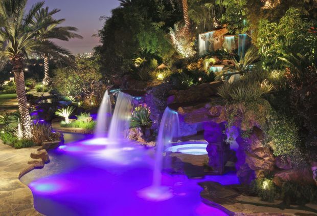 A dramatic tropical landscaped pool area at dusk, featuring a lagoon-style swimming pool with illuminated purple-blue underwater lighting, multiple cascading waterfalls, natural rock formations, dense palms and lush planting, and layered garden lighting. The scene has a resort-like, immersive outdoor atmosphere.