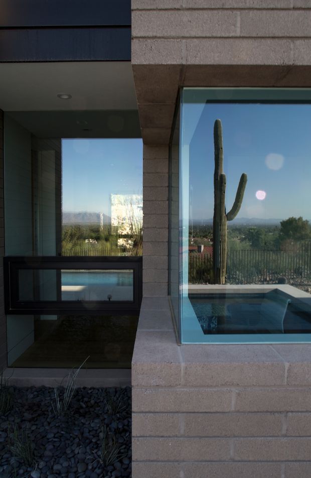 Exterior architectural detail of a modern masonry house featuring a projecting glazed window, dark metal framing, and a reflective pool or water feature integrated beside the facade. The glazing reflects a tall desert cactus, landscaped grounds, distant mountains, and clear blue sky. Dark gravel landscaping and recessed exterior lighting reinforce the minimalist desert-modern character.
