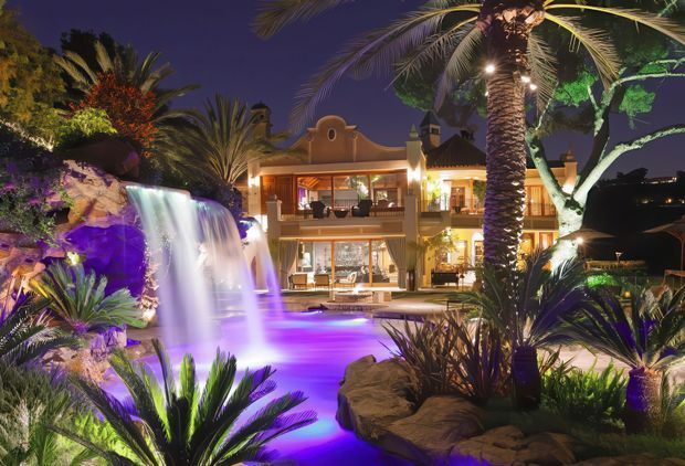 Night-time view of a lavish tropical resort-style property centered on a brightly illuminated swimming pool and waterfall feature. Dense palm planting, cycads, rocks, and dramatic purple and blue underwater or landscape lighting surround a multi-storey villa with balconies and warm interior lighting.