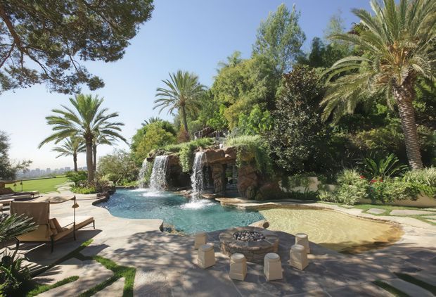 A luxurious landscaped outdoor pool area surrounded by mature tropical and subtropical planting, including palms and dense greenery. The pool features rock-clad waterfall cascades and a shallow adjoining section, with sun loungers, umbrellas, paved terraces, and a low outdoor seating arrangement around a fire feature. A manicured lawn and distant urban skyline are visible beyond the pool.
