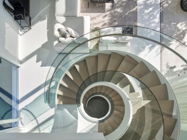 Top-down view of a sculptural spiral staircase with broad timber-toned treads, a dark central opening, and curved glass balustrades. The staircase occupies a bright, open interior void, with white walls, extensive glazing, and glimpses of pale seating and tiled flooring. Strong geometric shadows and natural daylight create a dramatic architectural composition.