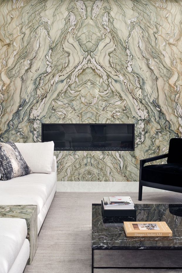 A refined living room featuring a dramatic bookmatched green-grey and cream marble feature wall with an integrated dark fireplace or television recess. A white upholstered sofa, black accent chair, dark marble coffee table, and neutral textured carpet create a luxurious, high-contrast interior.