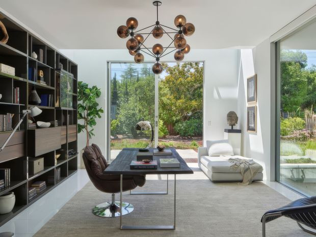 Bright, sophisticated living room or study with floor-to-ceiling glazing opening onto a landscaped garden. A dark built-in shelving wall filled with books and objects lines the left side, while a compact dark desk, leather swivel chair, white lounge chair, and ottoman occupy the room. A sculptural bronze globe pendant hangs centrally, with pale carpet, white walls, and framed artwork creating a refined contemporary setting.
