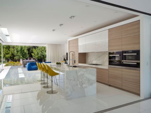 Bright, spacious contemporary open-plan kitchen with a large marble-look island, white waterfall sides, integrated sink and tap, and seating for four in mustard-yellow bar stools. Full-height timber-look and white cabinetry incorporates double wall ovens and a dark cooktop area, with a marble-look splashback. Glossy white tiled flooring reflects abundant daylight from extensive glazing and open access to a landscaped outdoor area. The room has a clean, luxurious, highly polished appearance.