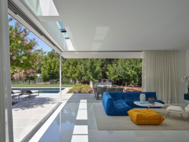 Bright contemporary living room opening through extensive floor-to-ceiling glazing to a pool terrace and landscaped garden. The room has a glossy pale tiled floor, white ceiling with large skylights or roof glazing, sheer white curtains, a sculptural vivid blue sectional sofa, mustard ottoman, and a low round coffee table. The open indoor-outdoor arrangement and strong natural light create an airy, refined setting.