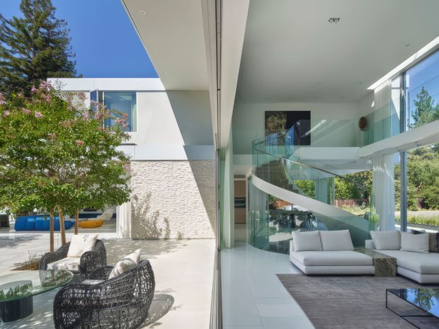 A bright contemporary residence photographed from a covered outdoor area, looking through full-height glazing into an open-plan living room. The interior features a dramatic curved glass balustrade and sculptural white staircase, pale tiled flooring, white walls, large windows, and light upholstered sofas. Outside, a paved courtyard contains dark woven lounge chairs, a glass-topped table, and landscaped trees. The architecture is clean-lined, highly glazed, and strongly connected to the courtyard.