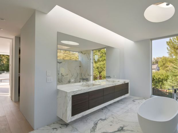 A bright, contemporary bathroom featuring a double vanity with dark wood cabinetry, a white marble benchtop and splashback, and matching marble floor tiles. A freestanding white bathtub sits beside large windows overlooking greenery. A full-width mirror, recessed ceiling lighting, and clean white walls create an airy, refined space.