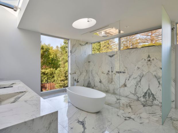 Bright contemporary bathroom featuring a freestanding white oval bathtub, expansive white veined marble wall and floor surfaces, a marble vanity, frameless glass shower enclosure, recessed ceiling lighting, and large windows overlooking trees.