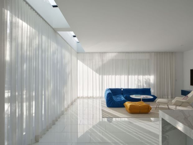 Bright, minimalist open-plan living room with floor-to-ceiling sheer curtains, glossy pale flooring, extensive skylights, and strong sunlight creating linear shadows. A vivid blue curved sofa and mustard ottoman sit around a small round table, with white lounge chairs and a marble-look kitchen island or counter in the foreground.