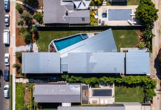 Aerial top-down view of a contemporary residential property in a suburban setting. The image is dominated by a long, light-coloured metal-roofed building with multiple roof forms, surrounding lawns, landscaped gardens, paved paths, and a bright blue swimming pool in the rear yard. Neighbouring homes and parked cars are visible around the site.