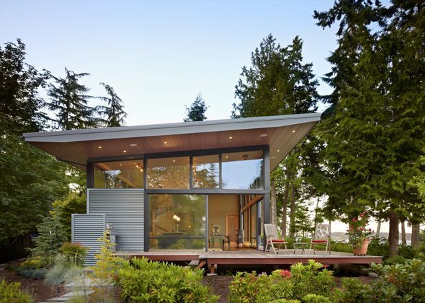 Exterior view of a compact modern pavilion-like home set within mature landscaped gardens. The building features extensive floor-to-ceiling glazing, a broad low-pitched roof with deep eaves, a warm timber-lined soffit with recessed lighting, corrugated metal wall cladding, and a raised timber deck with outdoor seating. Dense trees and planting surround the structure.