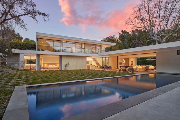 Contemporary two-storey residence with extensive floor-to-ceiling glazing, clean horizontal lines, and a sheltered outdoor living area arranged around a large rectangular swimming pool. Warm interior lighting glows through the glass at dusk, with landscaped grounds and mature trees surrounding the home.