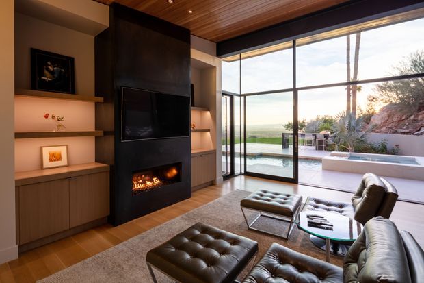 A refined contemporary living room with a dark built-in fireplace and television wall, warm timber ceiling, integrated wood cabinetry and illuminated display shelving. Large floor-to-ceiling glazed doors open the room to a paved outdoor terrace and swimming pool, with a distant coastal or open landscape view. Leather lounge seating, tufted ottomans and a low coffee table create a luxurious, comfortable arrangement.