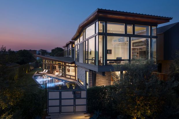 Twilight exterior view of a contemporary multi-level residence on a sloping site. The home features extensive floor-to-ceiling glazing, dark metal-framed windows, projecting upper-level volumes, horizontal rooflines, and warm illuminated interiors. A swimming pool and landscaped outdoor area are visible beside the house, with a garage or screened entry structure in the foreground.
