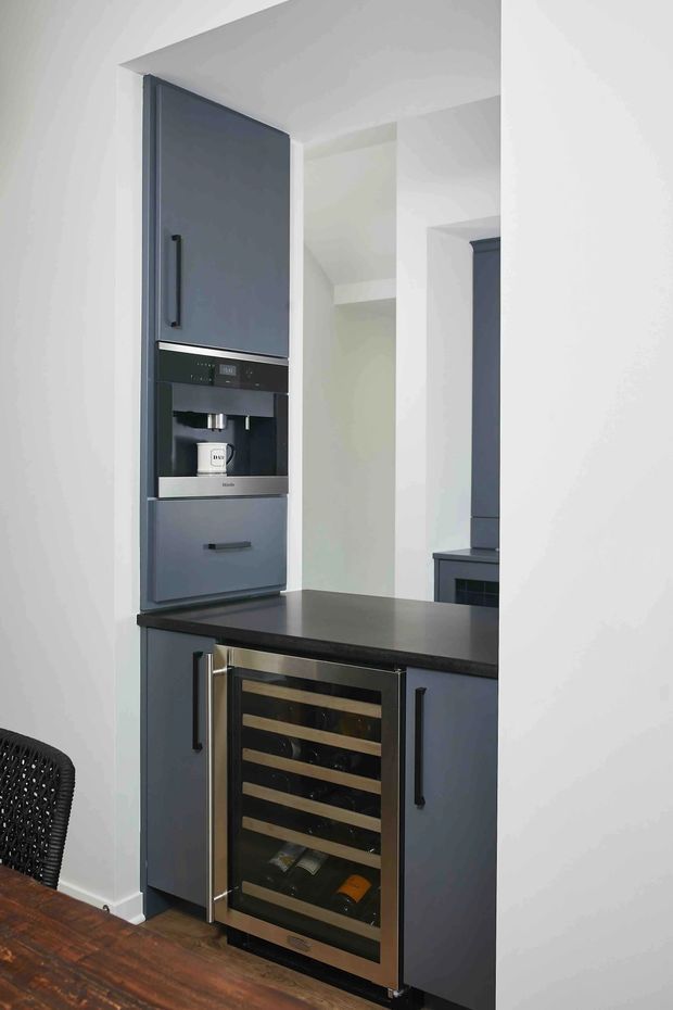 A compact built-in kitchen beverage station featuring dark blue-grey cabinetry, integrated coffee machine, black stone-like benchtop, and a stainless-framed glass-front wine refrigerator. White walls and dark timber flooring create a restrained, contemporary look. A partial dining table and chair are visible in the foreground.