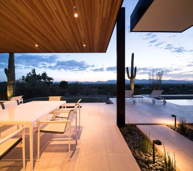 A refined outdoor entertaining terrace at dusk, with a large dining table and chairs beneath a warm timber-lined roof canopy. The tiled terrace extends toward a swimming pool and landscaped gravel garden, framed by dark structural posts and overlooking a wide desert-like suburban landscape with sculptural cacti. Integrated lighting creates a warm, inviting atmosphere against the cool evening sky.