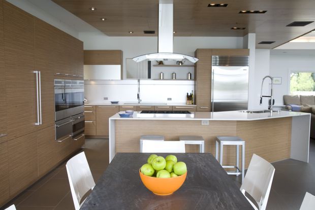 Open-plan contemporary kitchen and dining area with extensive light timber cabinetry, integrated stainless-steel appliances, a large island with white benchtops, and a dark dining table in the foreground. A sculptural stainless-steel rangehood, pendant-like ceiling detailing, and a living area visible beyond the kitchen contribute to the polished modern fit-out.