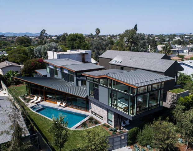 Aerial view of a large contemporary multi-level residence set within a leafy suburban neighbourhood. The home has several intersecting low-pitched metal roof forms, extensive floor-to-ceiling glazing, dark grey cladding and masonry walls with warm timber-toned trims. A long rectangular swimming pool, paved terrace and outdoor seating occupy the landscaped rear courtyard.